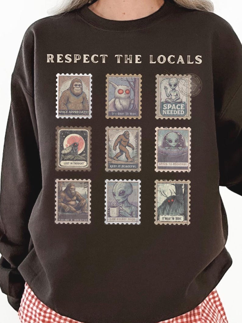 Cozy cryptid sweatshirt with a grid of vintage postage stamp illustrations including Mothman, Bigfoot, and alien folklore characters shown on a model wearing a Gildan 18000 Dark Chocolate crewneck.