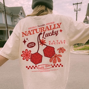 May include: Cream-colored t-shirt with a graphic design featuring the text "NATURALLY LUCKY" and two red dice. The design includes floral elements and the text "Critical Success!" and "Lucky Roller".