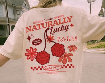 May include: Cream-colored t-shirt with a graphic design featuring the text "NATURALLY LUCKY" and two red dice. The design includes floral elements and the text "Critical Success!" and "Lucky Roller".