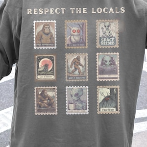 Cozy cryptid sweatshirt with a grid of vintage postage stamp illustrations including Mothman, Bigfoot, and alien folklore characters shown on a model wearing a Comfort Colors 1566 Pepper crewneck.