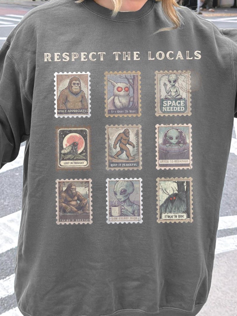 Cozy cryptid sweatshirt with a grid of vintage postage stamp illustrations including Mothman, Bigfoot, and alien folklore characters shown on a model wearing a Comfort Colors 1566 Pepper crewneck.