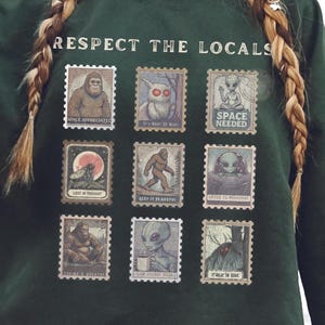 Cozy cryptid sweatshirt with a grid of vintage postage stamp illustrations including Mothman, Bigfoot, and alien folklore characters shown on a model wearing a Gildan 18000 Forest Green crewneck.