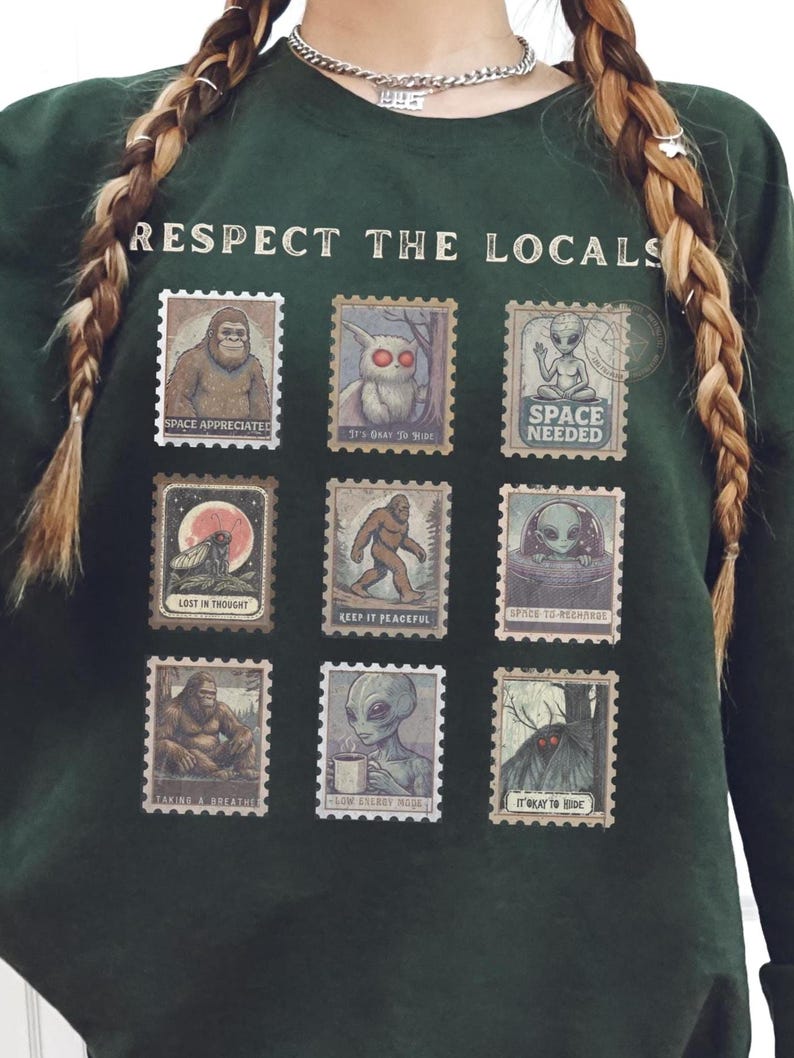 Cozy cryptid sweatshirt with a grid of vintage postage stamp illustrations including Mothman, Bigfoot, and alien folklore characters shown on a model wearing a Gildan 18000 Forest Green crewneck.