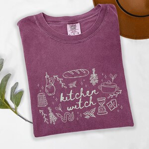 Kitchen Witch Comfort Colors t-shirt in Berry purple featuring a cottagecore witch design with bread loaf, hanging herbs, cooking pot, tarot cards, hourglass, snake, and botanical accents around the phrase "kitchen witch."