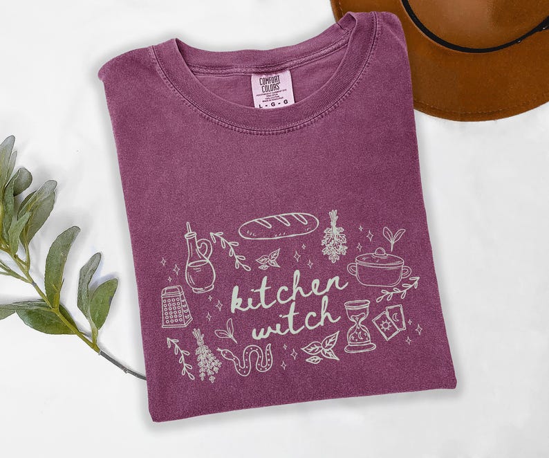 Kitchen Witch Comfort Colors t-shirt in Berry purple featuring a cottagecore witch design with bread loaf, hanging herbs, cooking pot, tarot cards, hourglass, snake, and botanical accents around the phrase "kitchen witch."