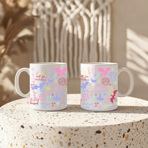 K&ouml;nnte beinhalten: Zwei wei&szlig;e Keramikbecher mit einem Fantasy-Motiv. Die Becher zeigen ein sich wiederholendes Muster aus Drachen, W&uuml;rfeln, Sternen und Text wie "roll for initiative" und "critical hit" in Pink, Lila und Blau. Die Becher stehen auf einer gesprenkelten Oberfl&auml;che.