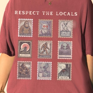 A Crimson colored Comfort Colors t-shirt with "Respect The Locals" cryptid stamp graphic, featuring Bigfoot, Mothman, and Alien stamps. Ideal for cryptid lovers and introverts.