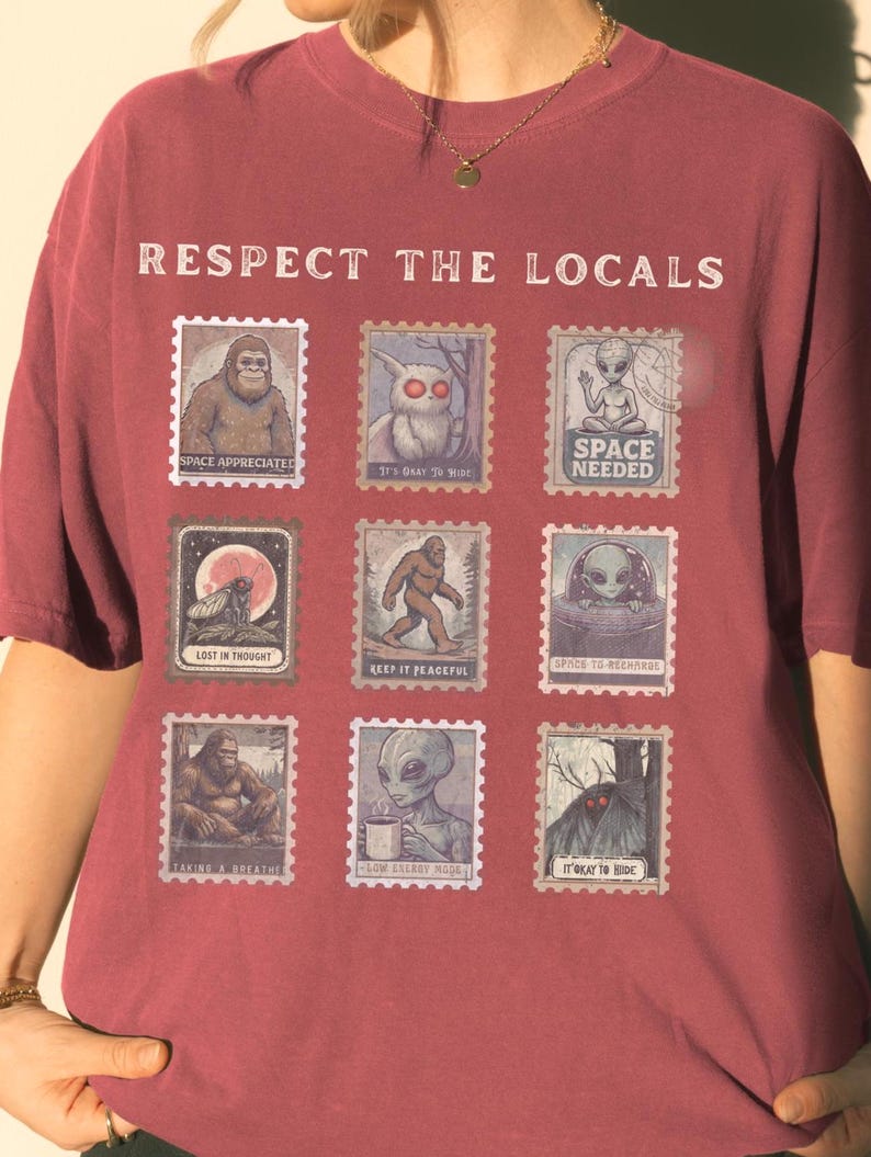A Crimson colored Comfort Colors t-shirt with "Respect The Locals" cryptid stamp graphic, featuring Bigfoot, Mothman, and Alien stamps. Ideal for cryptid lovers and introverts.