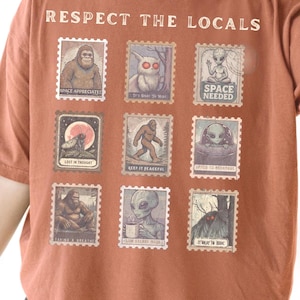 Yam colored Comfort Colors t-shirt with "Respect The Locals" cryptid stamp graphic, featuring Bigfoot, Mothman, and Alien stamps. Ideal for cryptid lovers and introverts.