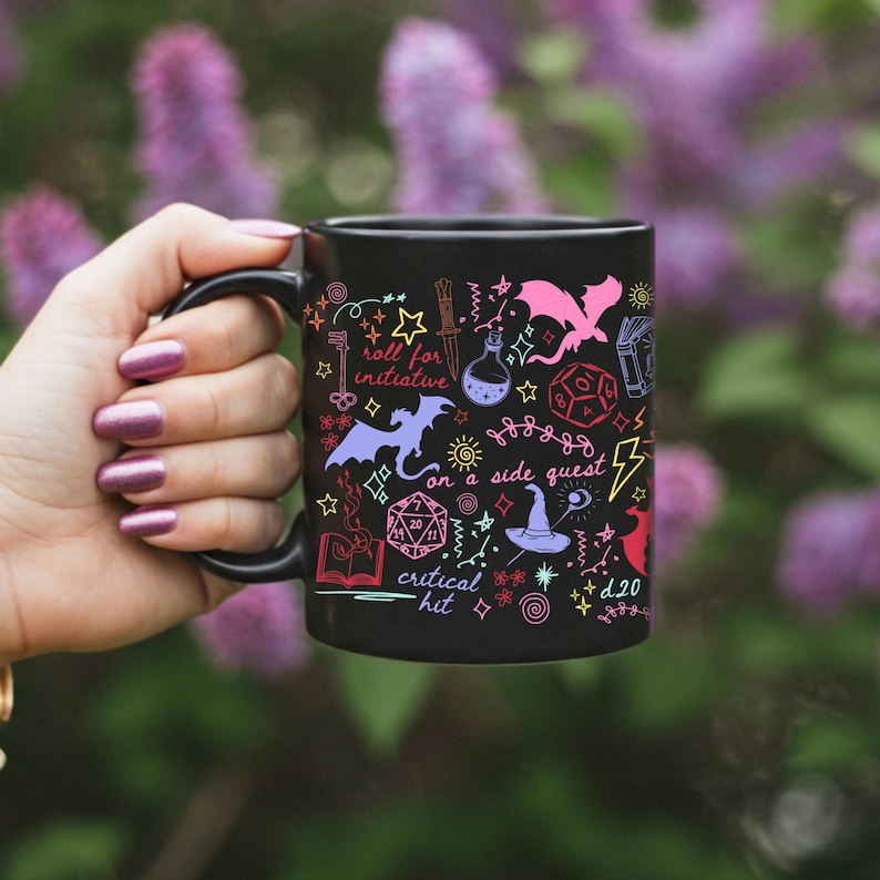 K&ouml;nnte beinhalten: Schwarze Keramik-Tasse mit farbenfrohen Fantasy-Illustrationen. Die Tasse zeigt einen Drachen, W&uuml;rfel, einen Zaubererhut und den Text "roll for initiative", "on a side quest" und "critical hit".