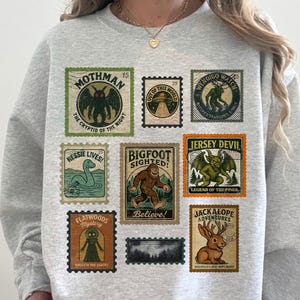 May include: A light gray crewneck sweatshirt featuring a graphic print of nine vintage-style postage stamps. The stamps depict various cryptids and legends, including Mothman, Bigfoot, the Jersey Devil, Nessie, and a Jackalope, with text labels.