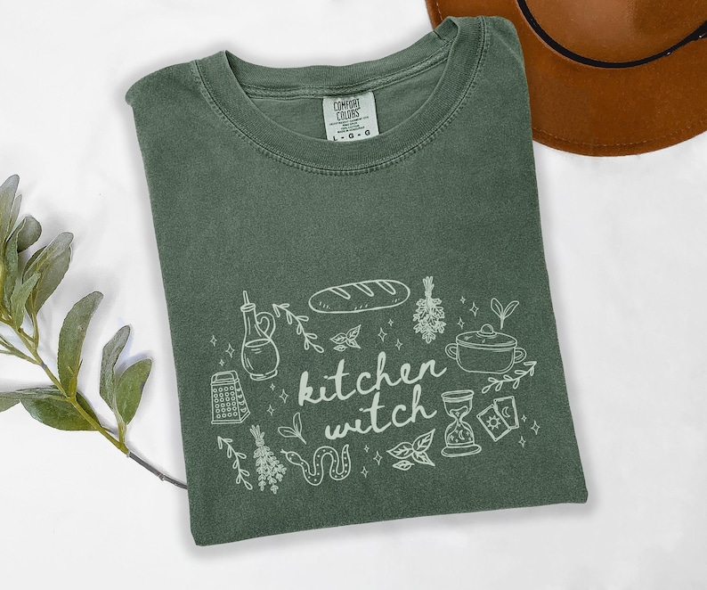 Kitchen Witch Comfort Colors t-shirt in Moss green featuring hand-drawn kitchen witch artwork with bread, herbs, cooking pot, grater, tarot cards, hourglass, snake, and botanical details.