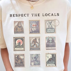 An Ivory colored Comfort Colors t-shirt with "Respect The Locals" cryptid stamp graphic, featuring Bigfoot, Mothman, and Alien stamps. Ideal for cryptid lovers and introverts.