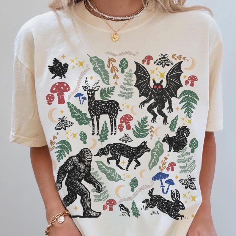 An Ivory Comfort Colors t-shirt featuring a forest-themed linocut cryptid design with Mothman, Bigfoot, and forest animals surrounded by mushrooms, ferns, stars, and moons in vintage woodland colors.