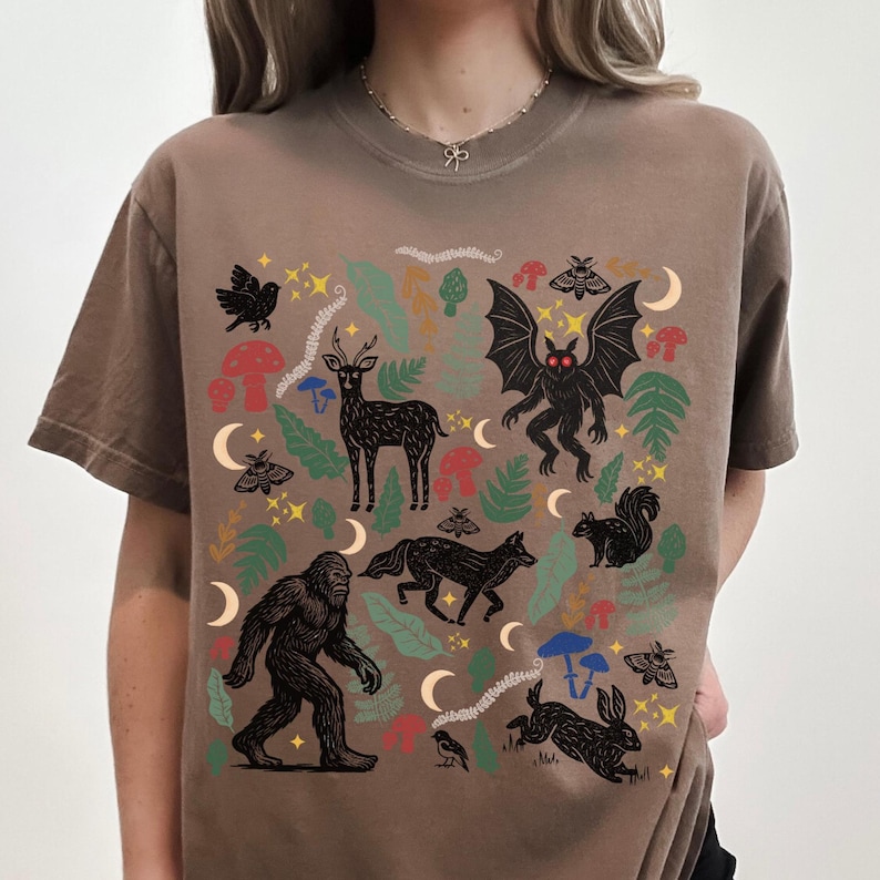 An Espresso Comfort Colors t-shirt featuring a forest-themed linocut cryptid design with Mothman, Bigfoot, and forest animals surrounded by mushrooms, ferns, stars, and moons in vintage woodland colors.