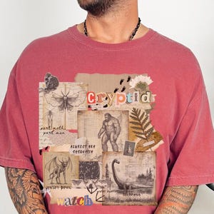 A man wears a Comfort Colors Crimson t shirt with a Cryptozoology design featuring Mothman, Bigfoot, Lochness and the Jersey Devil in a vintage field journal style. Intended for camping, hiking and outdoorsy activities or for lovers of spooky folklore.
