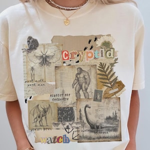 A Woman wears a Comfort Colors Ivory t shirt with a Cryptozoology design featuring Mothman, Bigfoot, Lochness and the Jersey Devil in a vintage field journal style. Intended for camping, hiking and outdoorsy activities or for lovers of spooky folklore.