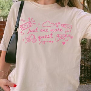 Woman wearing ivory cozy gamer shirt with pink just one more quest graphic in casual outdoor setting
