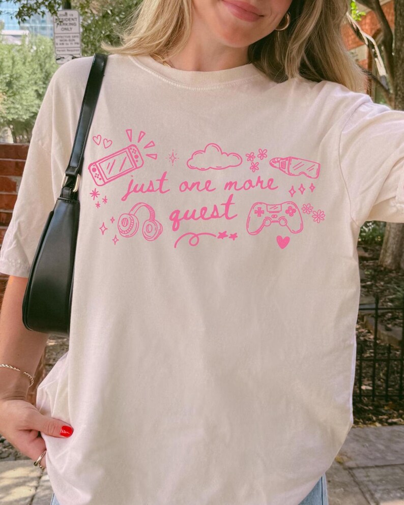 Woman wearing ivory cozy gamer shirt with pink just one more quest graphic in casual outdoor setting