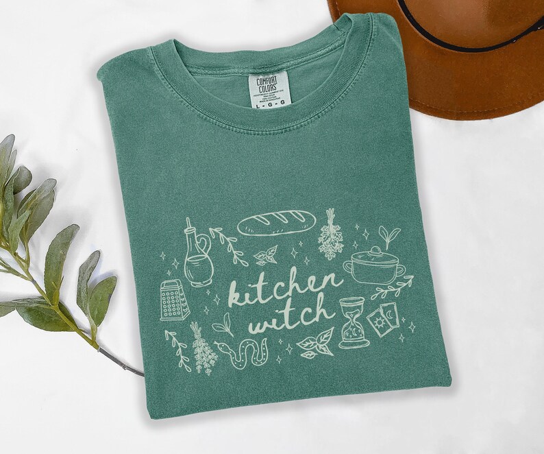 Kitchen Witch Comfort Colors t-shirt in Light Green featuring a witchy kitchen design with bread loaf, olive oil bottle, herbs, cooking pot, tarot cards, snake, and botanical elements around the phrase "kitchen witch."
