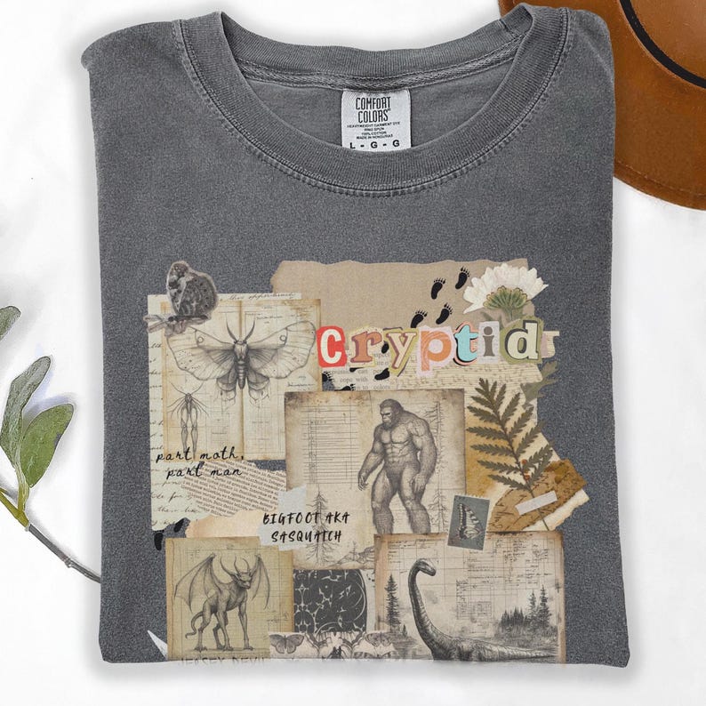 A folded Comfort Colors Pepper t shirt with a Cryptozoology design featuring Mothman, Bigfoot, Lochness and the Jersey Devil in a vintage field journal style. Intended for camping, hiking and outdoorsy activities or for lovers of spooky folklore.