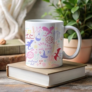 K&ouml;nnte beinhalten: Wei&szlig;e Keramiktasse mit farbenfrohen Fantasy-Illustrationen, darunter Drachen, W&uuml;rfel und Text mit der Aufschrift "roll for initiative", "on a side quest" und "critical hit". Die Tasse steht auf einem B&uuml;cherstapel.