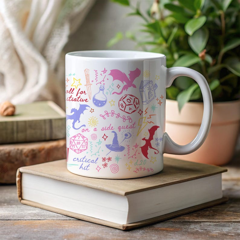 K&ouml;nnte beinhalten: Wei&szlig;e Keramiktasse mit farbenfrohen Fantasy-Illustrationen, darunter Drachen, W&uuml;rfel und Text mit der Aufschrift "roll for initiative", "on a side quest" und "critical hit". Die Tasse steht auf einem B&uuml;cherstapel.