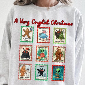 May include: A light gray crewneck sweatshirt featuring the red text A Very Cryptid Christmas above nine postage stamp-style illustrations of various cryptids and aliens in festive holiday scenes, including Bigfoot, Mothman, and extraterrestrials.