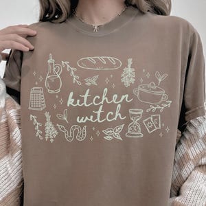 Woman wearing an Espresso brown Comfort Colors t-shirt featuring the phrase "kitchen witch" surrounded by hand-drawn bread, herbs, cooking tools, tarot cards, hourglass, snake, and botanical illustrations.