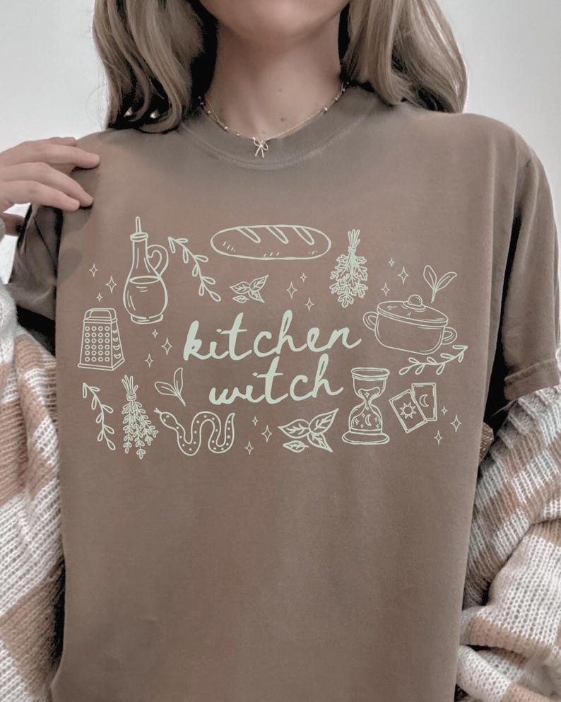 Woman wearing an Espresso brown Comfort Colors t-shirt featuring the phrase "kitchen witch" surrounded by hand-drawn bread, herbs, cooking tools, tarot cards, hourglass, snake, and botanical illustrations.