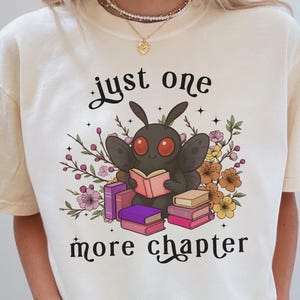 May include: A cream-colored t-shirt featuring a graphic of a black moth with red eyes reading a book, surrounded by colorful flowers and stacks of books. The text "just one more chapter" is printed in black script above and below the illustration.