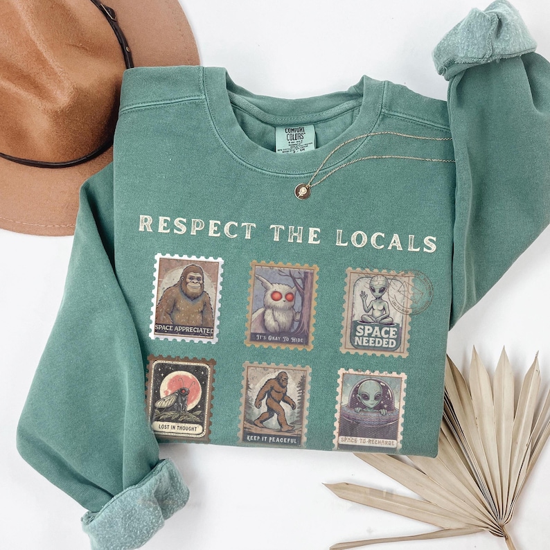 A folded flat lay of a Light Green colored Comfort Colors sweatshirt with "Respect The Locals" cryptid stamp graphic, featuring Bigfoot, Mothman, and Alien stamps. Ideal for cryptid lovers and introverts.