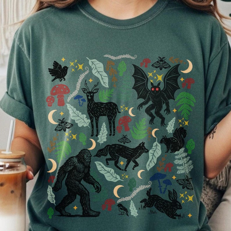 A Blue Spruce Comfort Colors t-shirt featuring a forest-themed linocut cryptid design with Mothman, Bigfoot, and forest animals surrounded by mushrooms, ferns, stars, and moons in vintage woodland colors.