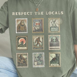 A moss colored Comfort Colors t-shirt with "Respect The Locals" cryptid stamp graphic, featuring Bigfoot, Mothman, and Alien stamps. Ideal for cryptid lovers and introverts.