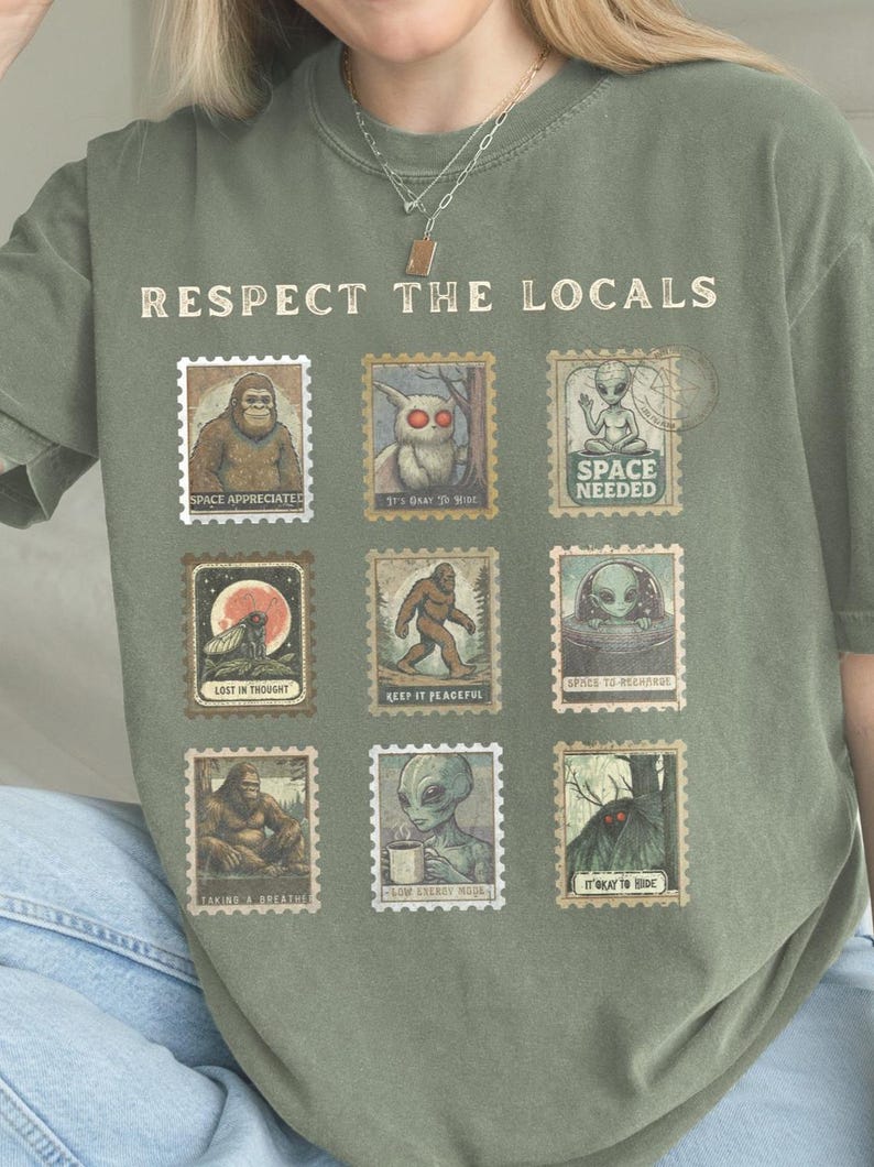 A moss colored Comfort Colors t-shirt with "Respect The Locals" cryptid stamp graphic, featuring Bigfoot, Mothman, and Alien stamps. Ideal for cryptid lovers and introverts.