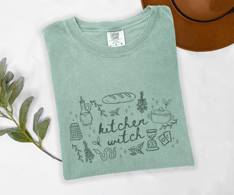 Kitchen Witch Comfort Colors t-shirt in Bay blue-green featuring hand-drawn bread, herbs, cooking pot, olive oil bottle, tarot cards, hourglass, snake, and botanical illustrations surrounding the words "kitchen witch."