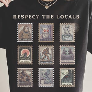 A black Comfort Colors t-shirt with "Respect The Locals" cryptid stamp graphic, featuring Bigfoot, Mothman, and Alien stamps. Ideal for cryptid lovers and introverts.