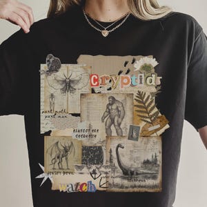 A Woman wears a Comfort Colors Black t shirt with a Cryptozoology design featuring Mothman, Bigfoot, Lochness and the Jersey Devil in a vintage field journal style. Intended for camping, hiking and outdoorsy activities or for lovers of spooky folklore.
