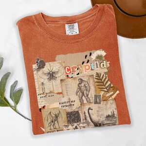 A folded Comfort Colors Yam t shirt with a Cryptozoology design featuring Mothman, Bigfoot, Lochness and the Jersey Devil in a vintage field journal style. Intended for camping, hiking and outdoorsy activities or for lovers of spooky folklore.