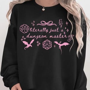May include: A black long-sleeve sweatshirt featuring a light pink graphic print. The design includes the text "literally just a dungeon master" surrounded by illustrations of two twenty-sided dice, two flying dragons, a bow, a lightning bolt, and stars.
