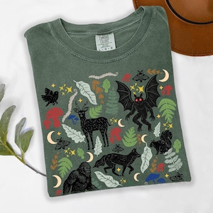 A Moss Comfort Colors t-shirt featuring a forest-themed linocut cryptid design with Mothman, Bigfoot, and forest animals surrounded by mushrooms, ferns, stars, and moons in vintage woodland colors.