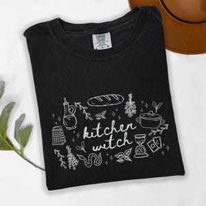 Kitchen Witch Comfort Colors t-shirt in Black with cream-colored artwork featuring bread, herbs, cooking tools, tarot cards, hourglass, snake, and botanical illustrations surrounding the words "kitchen witch."