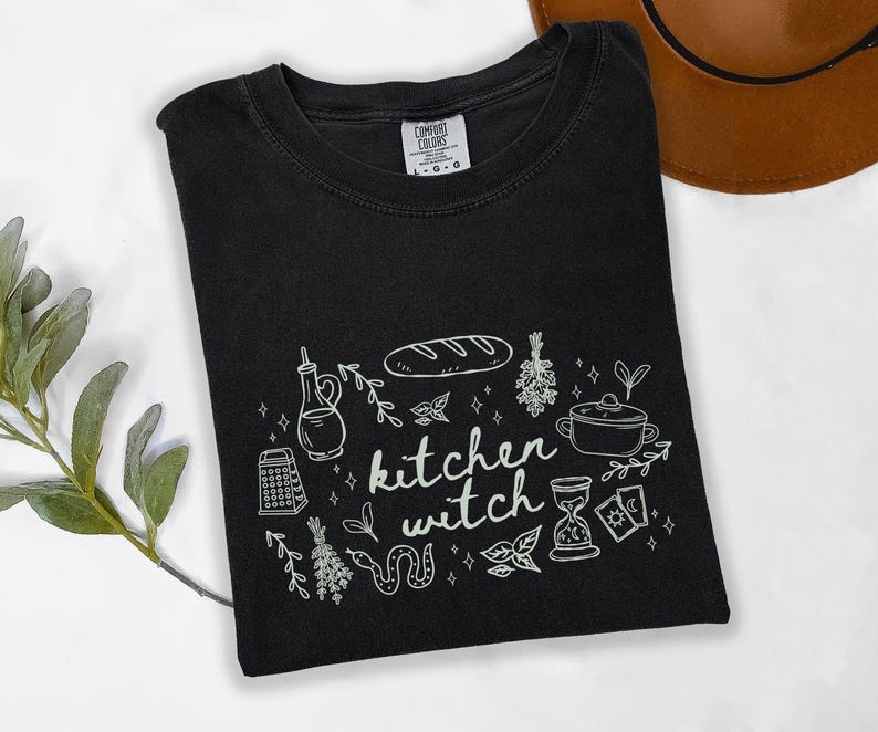 Kitchen Witch Comfort Colors t-shirt in Black with cream-colored artwork featuring bread, herbs, cooking tools, tarot cards, hourglass, snake, and botanical illustrations surrounding the words "kitchen witch."
