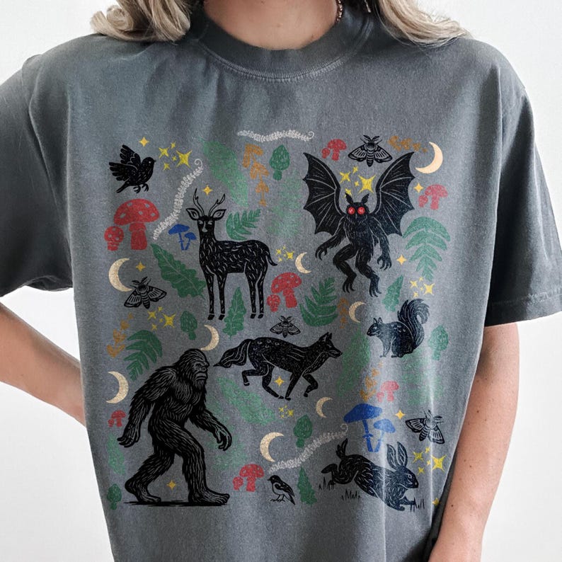 A Pepper Comfort Colors t-shirt featuring a forest-themed linocut cryptid design with Mothman, Bigfoot, and forest animals surrounded by mushrooms, ferns, stars, and moons in vintage woodland colors.