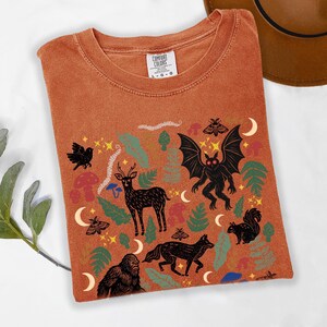 A Yam Comfort Colors t-shirt featuring a forest-themed linocut cryptid design with Mothman, Bigfoot, and forest animals surrounded by mushrooms, ferns, stars, and moons in vintage woodland colors.