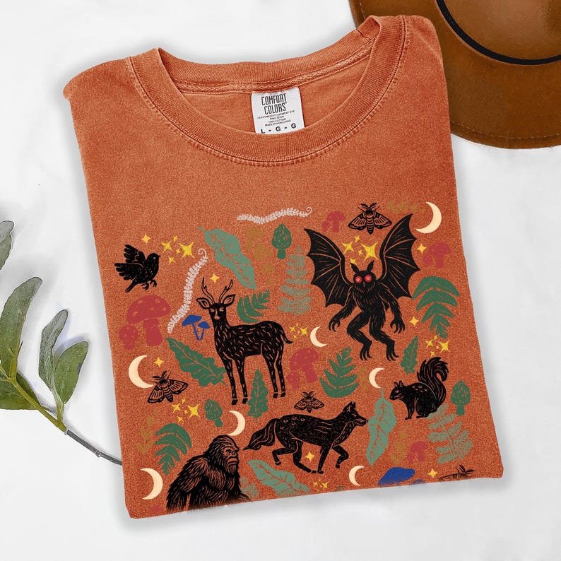 A Yam Comfort Colors t-shirt featuring a forest-themed linocut cryptid design with Mothman, Bigfoot, and forest animals surrounded by mushrooms, ferns, stars, and moons in vintage woodland colors.