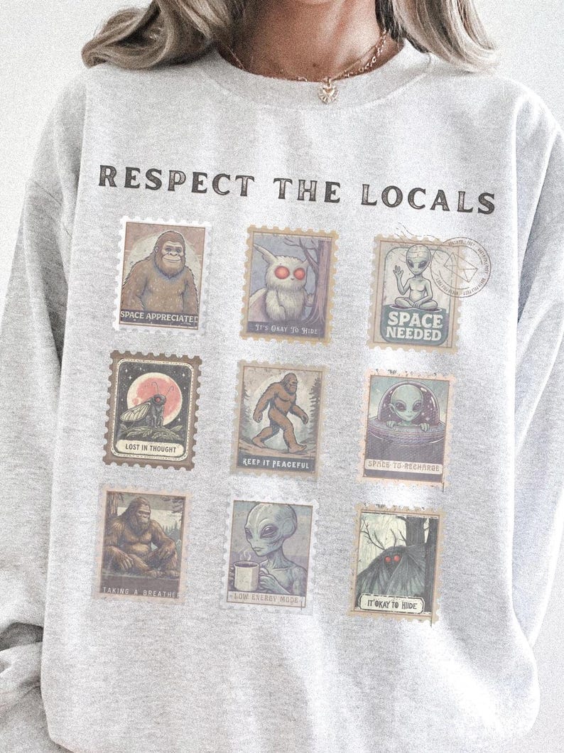 Cozy cryptid sweatshirt with a grid of vintage postage stamp illustrations including Mothman, Bigfoot, and alien folklore characters shown on a model wearing a Gildan 18000 Ash crewneck.