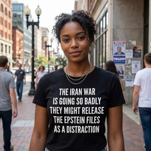 Puede incluir: Una camiseta negra con texto blanco: "THE IRAN WAR IS GOING SO BADLY THEY MIGHT RELEASE THE EPSTEIN FILES AS A DISTRACTION." La persona que lleva la camiseta está de pie en una acera de la ciudad.