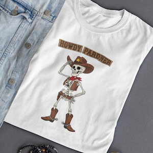 May include: White t-shirt with a cartoon skeleton cowboy design, complete with a brown hat, boots, and a red neckerchief. The text "HOWDY PARDNER" is above the skeleton. The shirt is shown with a denim jacket, sunglasses, and bracelets.