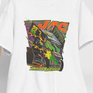May include: White t-shirt featuring a colorful graphic of a race car with the text "JAC" and "Hayden Schild". The design includes green, yellow, and black elements. A white baseball cap hangs on a wooden chair.